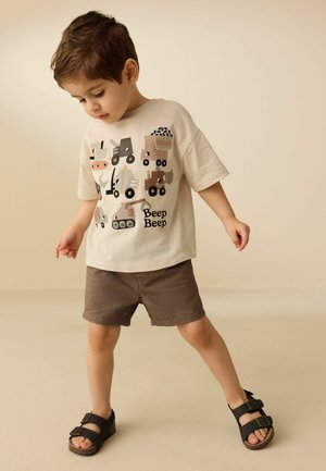 Young boy in beige construction vehicle print shirt, brown shorts, and black sandals, standing with legs apart on light background.