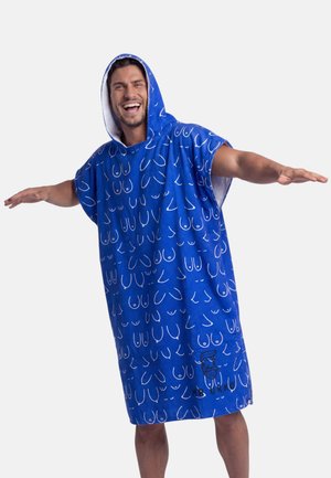 Hombre con un poncho de toalla azul con capucha y dibujos lineales blancos, sonriendo y extendiendo los brazos contra un fondo liso.