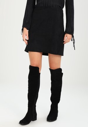 Jupe en suède noire à coupe droite, associée à des bottes en suède noires au-dessus du genou. La tenue est complétée par un haut à manches longues texturé.