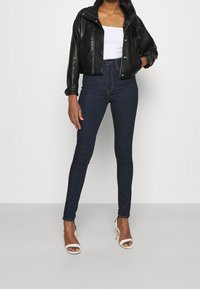 Chaqueta corta de cuero sintético negra, top ajustado blanco, jeans skinny de tiro alto azul oscuro y sandalias de tacón alto blancas con tiras.