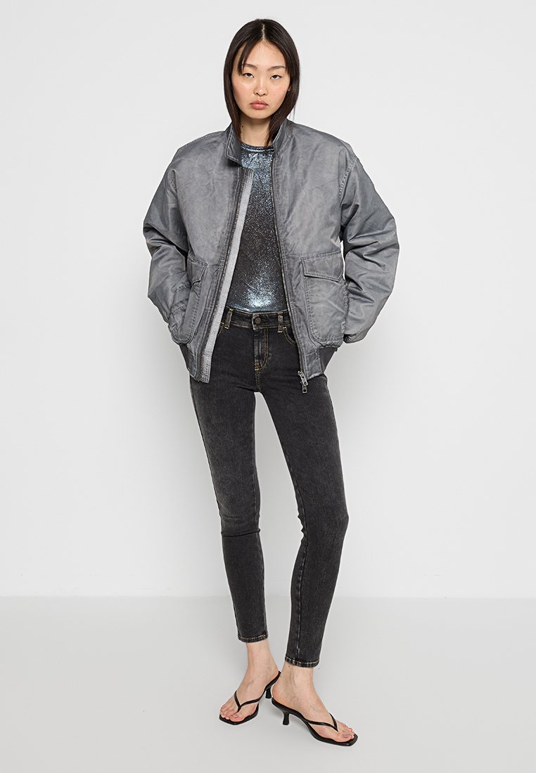 Giacca bomber grigia con zip frontale e due tasche, abbinata a un top blu scintillante e jeans skinny neri, completata da sandali neri.