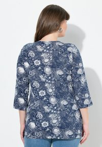 Frau mit braunen Haaren, die silberne Creolen trägt und ein navyblaues Langarmoberteil mit weißen spiralförmigen Blumenmustern anhat, steht vor einer weißen Wand.