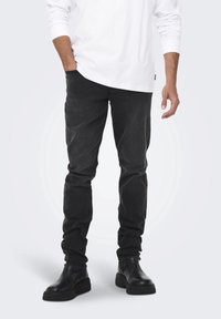 Jeans neri in denim con taglio slim, abbinati a una camicia bianca lunga e stivaletti neri. Il tessuto è liscio con un leggero effetto slavato.