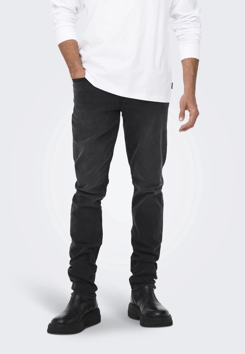 Jeans neri in denim con taglio slim, abbinati a una camicia bianca lunga e stivaletti neri. Il tessuto è liscio con un leggero effetto slavato.