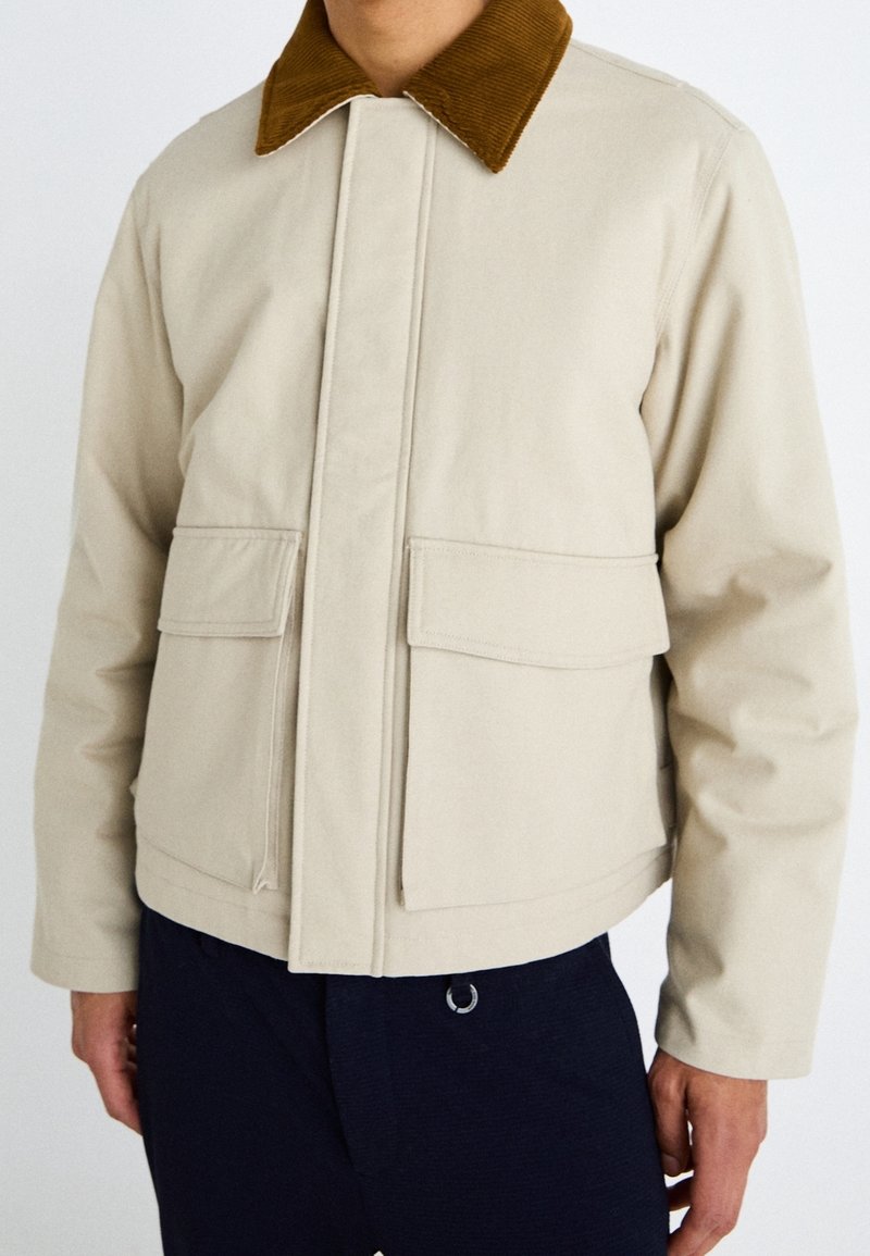 Chaqueta aislante beige con un cuello de pana marrón, bolsillos frontales con solapa y cierre de cremallera, que presenta un diseño corto y una textura suave.