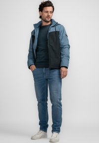 Chaqueta con cremallera azul y negra con capucha, que presenta una textura suave y paneles en contraste; llevada con un suéter oscuro, jeans y zapatillas blancas.