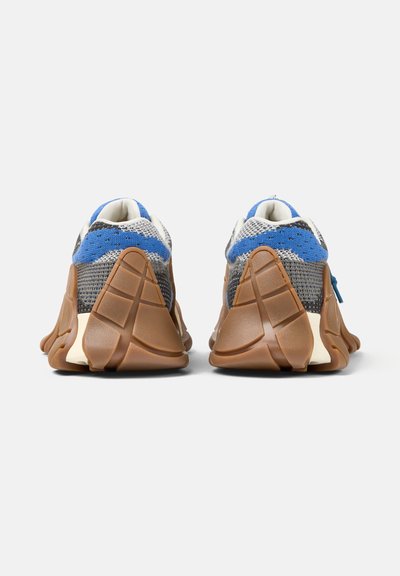 Zapatillas deportivas con partes superiores de punto en azul, gris y blanco, y suelas gruesas de goma marrón segmentadas, vistas desde atrás sobre un fondo blanco.