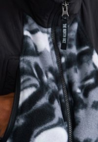 Chaqueta negra con cremallera y forro de forro polar gris y blanco con patrón. Presenta un tirador de cremallera etiquetado y tejido texturizado.