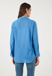 Camicia blu a maniche lunghe realizzata in tessuto liscio, con colletto classico, spalle basse, yoke posteriore e polsini con bottoni.