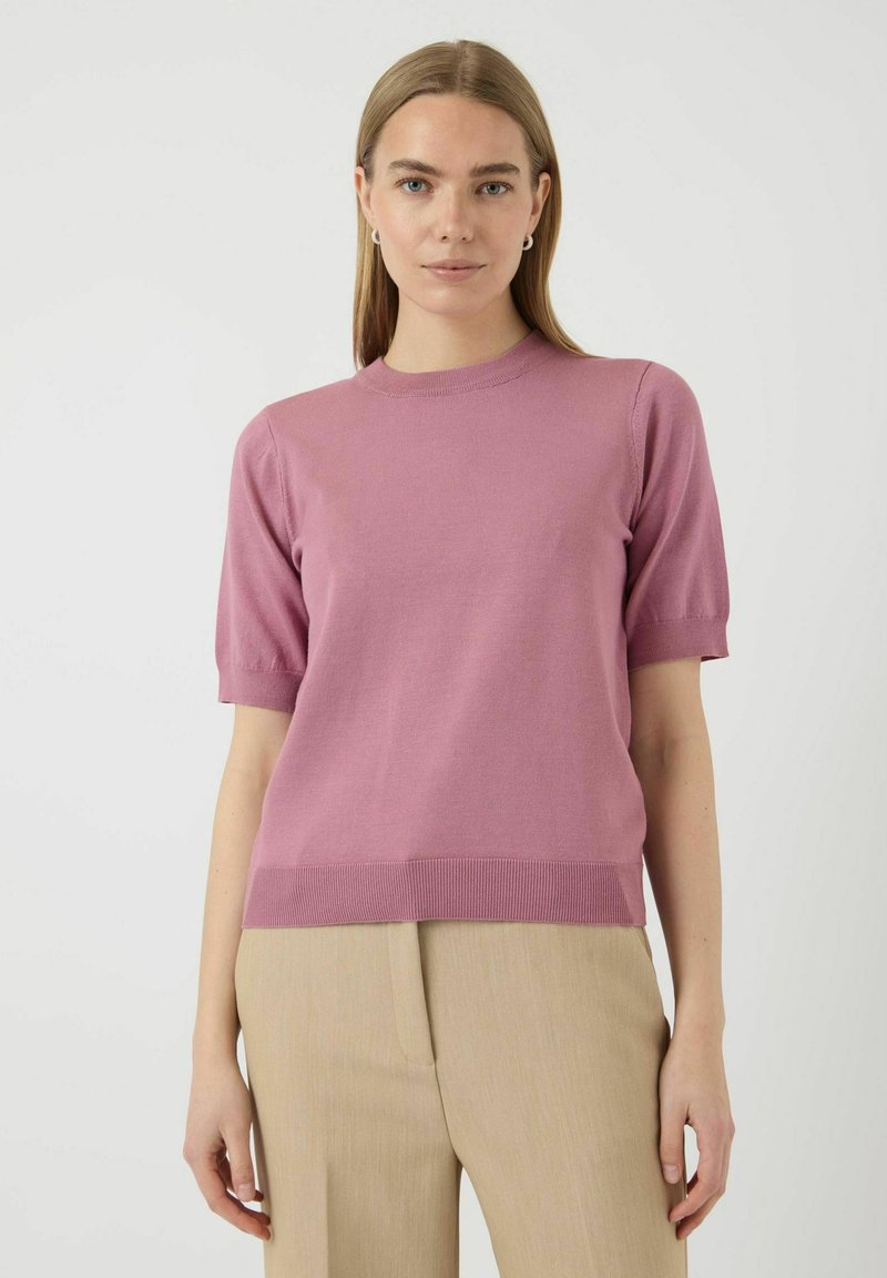 Svijetloružičasti pleteni pulover s kratkim rukavima, okruglog izreza i rebrastim rubom, uparen s svijetlobijelim krojenim hlačama.