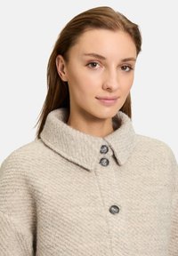 Manteau en laine beige avec un motif texturé, grand col et trois grands boutons sombres à l'avant. Tissu doux avec une coupe décontractée.