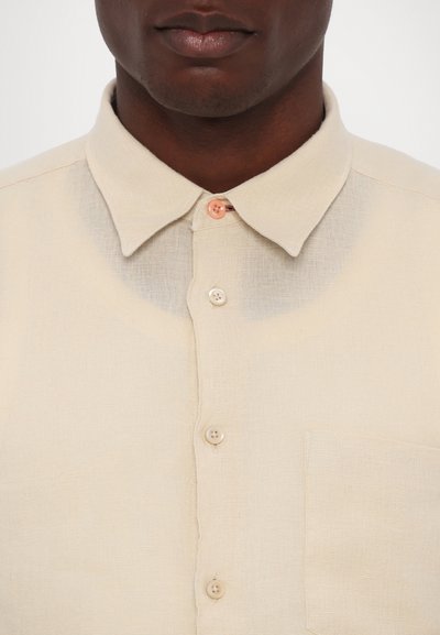Camicia beige con colletto, in tessuto testurizzato, caratterizzata da cinque bottoni, una tasca e una leggera variazione di colore vicino al colletto.