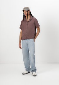 Kortærmet, løs pasform brunternet skjorte med knapper, parret med lyseblå jeans og en grå kasket med logo, båret med sort og hvid sneakers.