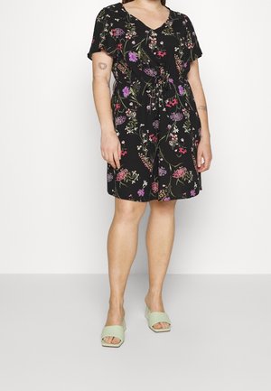 Osoba w czarnej sukience w kwiaty i jasnozielonych sandałach na blokowym obcasie z odkrytymi palcami, stojąca na tle jasnego, jednolitego tła.