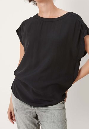 Persoon die een losse zwarte korte mouw blouse en lichtgrijze spijkerbroek draagt met één hand in de zak tegen een effen achtergrond.