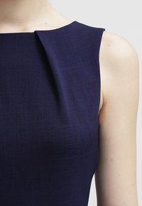 Mekko, joka on tummansininen ilman hihoja, ja jossa on muotoiltu pääntie sekä hienovaraisia laskoksia vartalon alueella. Sujuva tekstuuri ja tiukka muotoilu.