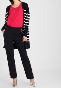 Navy and white striped cardigan over a red pocket t-shirt; black trousers with side stripes; silver high-heeled shoes.