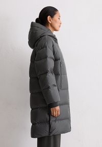 Graue gepolsterte Winterjacke mit Kapuze, ausgestattet mit einer Reißverschlusstasche und gerippten Bündchen, die eine weiche, steppartige Textur zeigt.