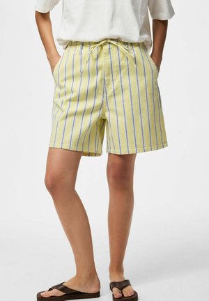 Personne portant un short jaune à rayures bleues verticales avec un cordon, associé à un haut blanc et des tongs marron. Mains dans les poches.