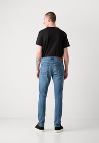 Jeans de mezclilla azul con corte recto, que cuentan con un parche de cuero marrón en la parte trasera, combinados con una camiseta negra y zapatos negros.