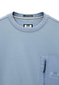 Camisa de cuello redondo en azul claro con un bolsillo en el pecho con cremallera y etiqueta de "Weekend Offender", además de una etiqueta en el cuello que dice "Admit Nothing".