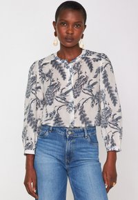 Donna che indossa una camicia bianca a bottoni con stampa di pavoni blu e foglie, abbinata a jeans blu a vita alta e grandi orecchini a goccia.