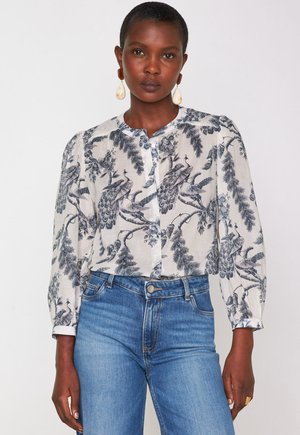 Donna che indossa una camicia bianca a bottoni con stampa di pavoni blu e foglie, abbinata a jeans blu a vita alta e grandi orecchini a goccia.