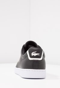 Svarta lädersneakers med en vit gummisula. Funktioner inkluderar en texturerad svart tygtunga och en vit krokodil-logotyp på hälen.