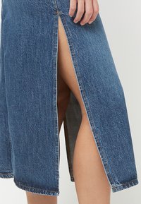 Mână sprijinită pe o fustă de denim albastră până la genunchi, cu o fantă care lasă să se vadă partea superioară a piciorului, pe un fundal neutru.