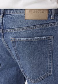 Traperice u svijetloplavom pranju s kožnom oznakom na leđima sa utisnutim natpisom "ONLY & SONS". Detalji uključuju stražnju vrećicu i naglašene šavove.