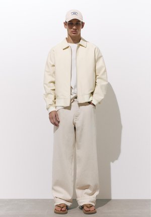 Fiatal férfi krémszínű dzsekiben, széles krémszínű nadrágban, bézs sapkában és barna papucsban, fehér fal előtt áll.