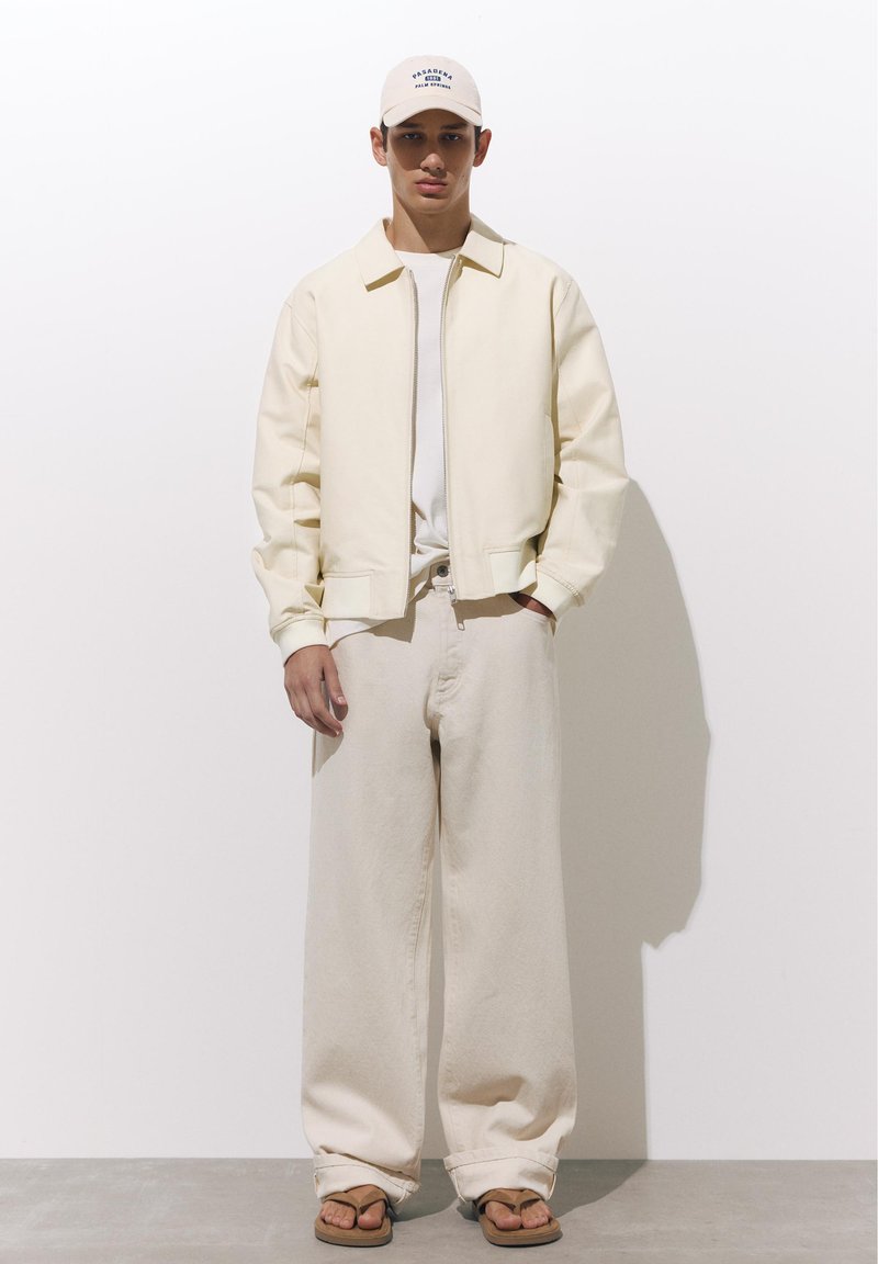 Jeune homme portant une veste crème, un pantalon large crème, une casquette beige et des tongs marron, debout contre un mur blanc.
