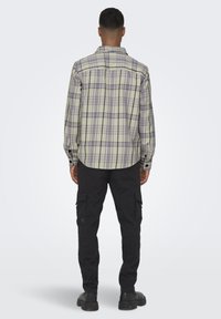 Long-sleeve plaid shirt in light green and purple, featuring a button-down collar and rear yoke; paired with black cargo pants and boots.