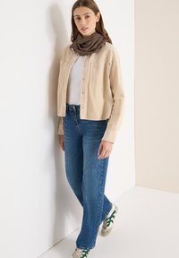 Veste en velours côtelé beige à coupe décontractée, associée à un jean bleu clair à jambes droites. Une écharpe marron et des baskets avec des touches vertes complètent le look.
