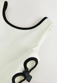 Vêtement blanc sans manches avec un bord noir autour de l'encolure et des emmanchures, comportant des nœuds circulaires noirs comme accents décoratifs.