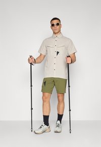 Beige shirt met korte mouwen en twee voorkruiken, groene korte broek, zwarte trekkingstokken en grijze sneakers. Zonnebril maakt de outfit compleet.
