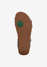 Ruskohvinen kumipohja herringbone-kuviolla, vihreä pyöreä logo yksityiskohtana ja tumma yläosa näkyvissä reunoilla.