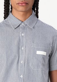 Chemise à manches courtes boutonnée à rayures bleu marine et blanches. Présente une poche poitrine avec une étiquette blanche et des coutures contrastées.