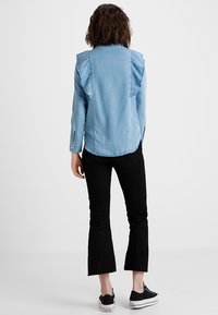 Camicia di denim azzurro chiaro con spalle arricciate, maniche lunghe e orlo curvo. Abbinata a pantaloni in velluto a coste neri svasati e sneakers nere.
