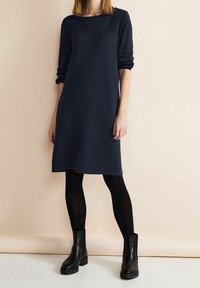 Person som bär en mörkblå långärmad klänning med svarta strumpbyxor och svarta ankelskor, stående mot en beige bakgrund.