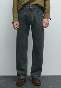 Persona che indossa jeans in denim scuro a taglio ampio, una camicia a maniche lunghe verde oliva e scarpe in suede marroni, in piedi su un pavimento grigio.