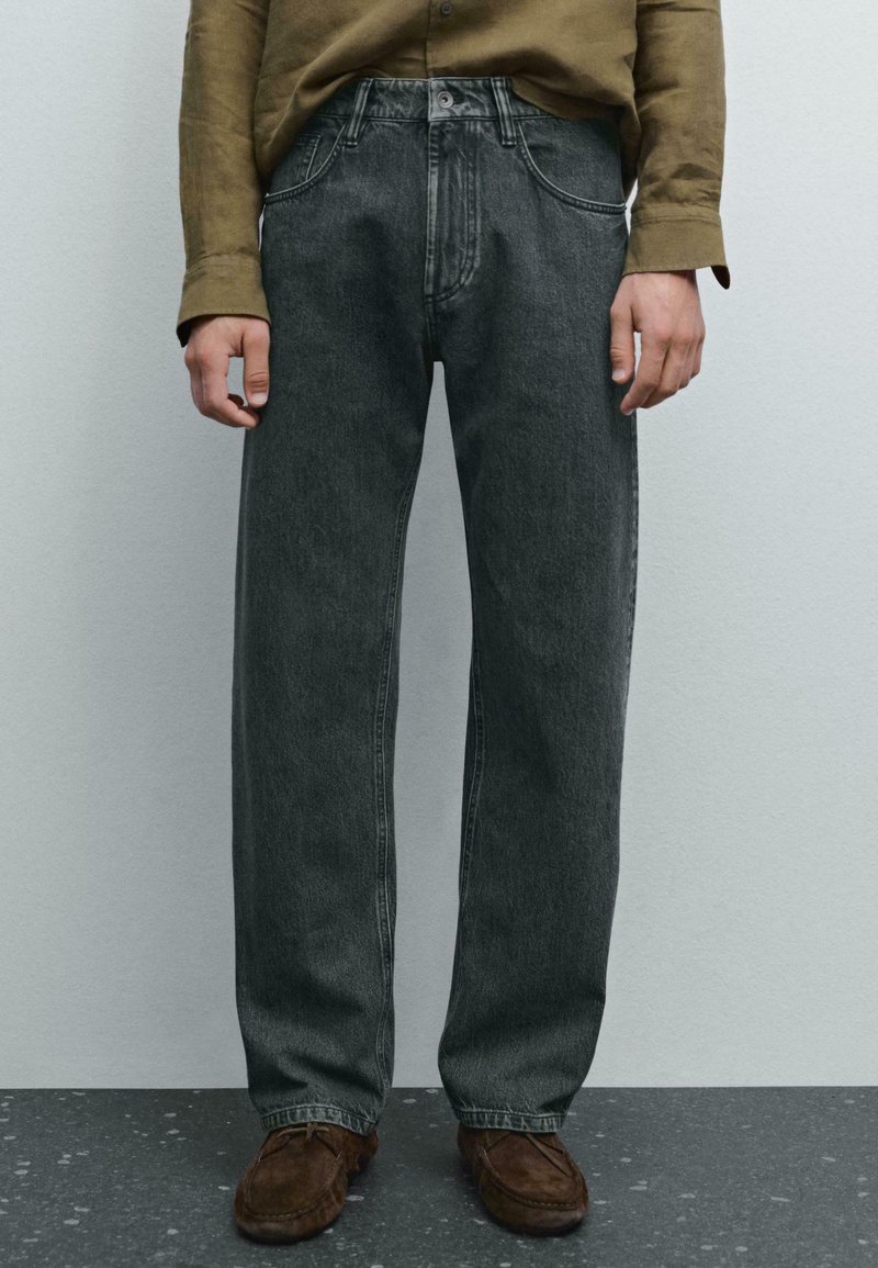 Persona che indossa jeans in denim scuro a taglio ampio, una camicia a maniche lunghe verde oliva e scarpe in suede marroni, in piedi su un pavimento grigio.