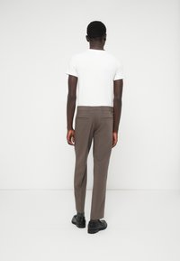 Pantaloni eleganti marroni con vestibilità slim, dotati di due tasche posteriori, abbinati a una t-shirt bianca a maniche corte e scarpe nere.