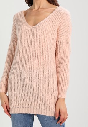 Mujer que lleva un suéter de punto grueso color rosa claro con cuello en V y mangas largas, junto con unos vaqueros azules.