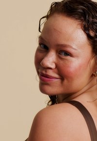 Bruine, gestructureerde haar, draagt een bruine spaghettiband top. Huid met sproeten, lichte blush. Zachte uitdrukking, zijaanzicht tegen een beige achtergrond.
