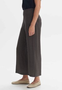 Kobieta w wysokich, szerokich spodniach o wzorze geometrycznym i beżowych balerinach, stojąca na białym tle.