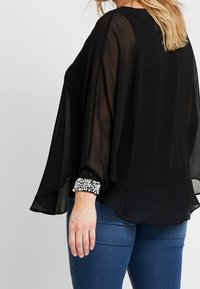 Blouse noire transparente avec des manches larges et un col rond. Des embellissements brillants ornent les poignets. Associée à un jean bleu.