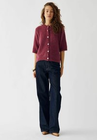 Fuzzy burgunder cardigan med korte ærmer og hvide knapper, parret med mørkeblå brede jeans. Glat strikket tekstur og afslappet pasform.