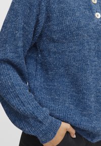 Pull en tricot bleu avec une finition texturée, présentant une coupe décontractée, des manches longues et deux grands boutons au col.