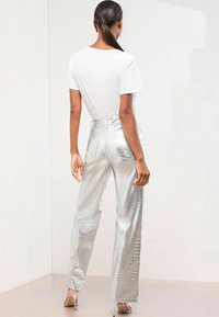 Srebrne, teksturirane široke hlače z bleščečim zaključkom, v kombinaciji z belo majico s kratkimi rokavi. Stoječi položaj, zadnji pogled.